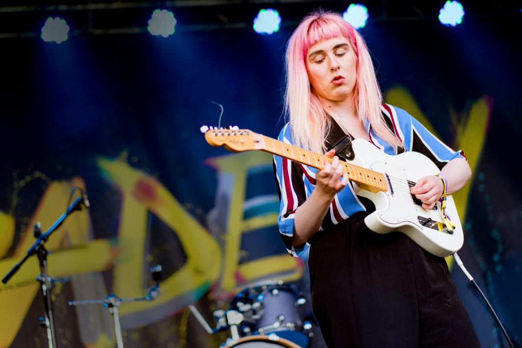Person with long, light-colored hair playing an electric guitar, wearing a striped shirt and black pants. Their face is blurred. Stage lights and part of a drum set are visible in the background.